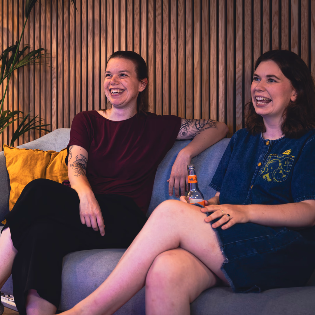 Zwei lachende junge Frauen mit Tattoos sitzen entspannt auf einem Sofa vor einer Holzlattenwand, eine hält eine Flasche Getränk.