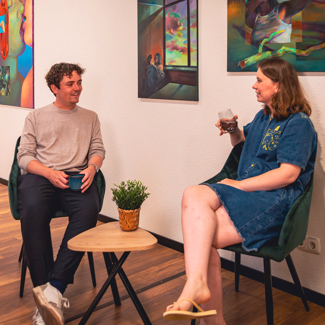 Zwei Menschen sitzen sich in einem modernen Raum gegenüber, trinken und unterhalten sich neben einem kleinen Holztisch mit einer Pflanze.
