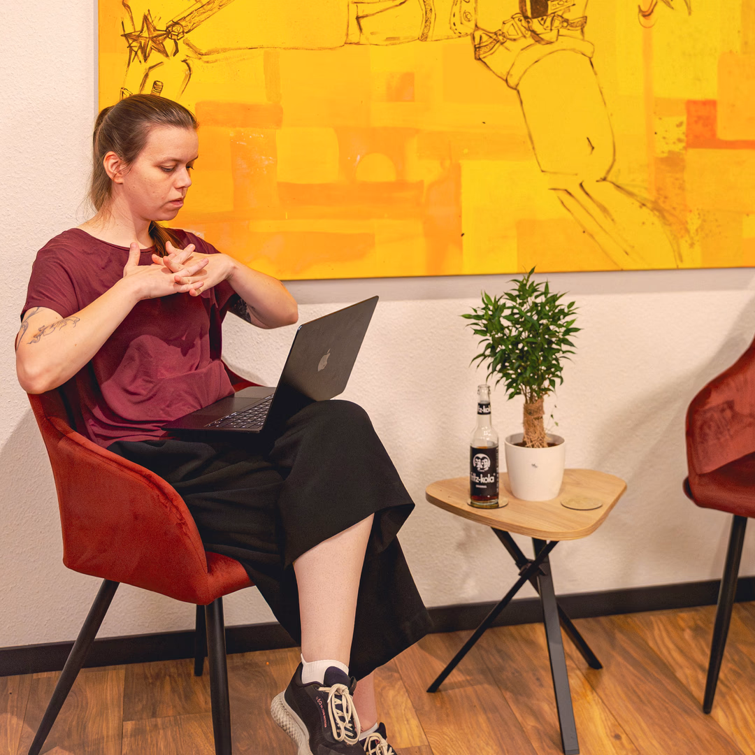 Frau sitzt in einem roten Stuhl mit einem Laptop auf dem Schoß, im Hintergrund ein gelbes Kunstwerk und ein kleiner Tisch mit einer Pflanze und einer Flasche.