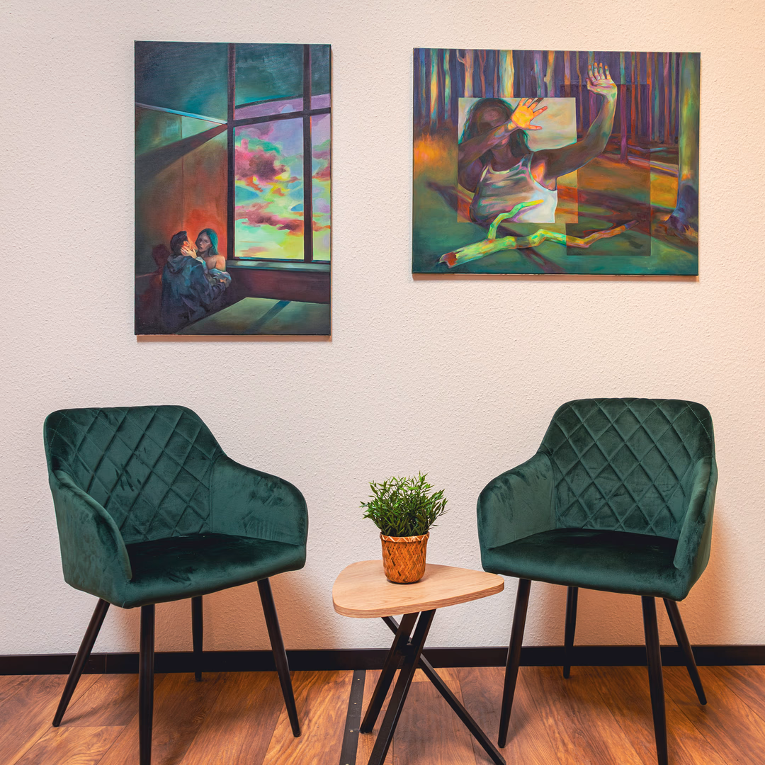 Zwei grüne Sessel mit zweieckigem Holz- und Metalltisch dazwischen, darauf ein Topf mit einer kleinen Pflanze; zwei farbenfrohe Gemälde hängen an der Wand.