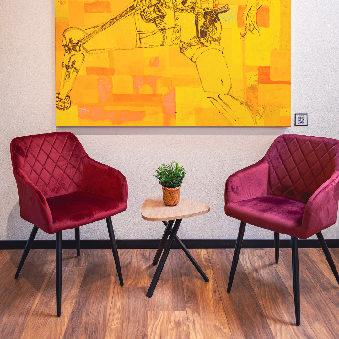 Zwei rote gepolsterte Stühle mit schwarzem Metallgestell um einen kleinen dreieckigen Holztisch mit einer kleinen Topfpflanze davor ein großes gelb-orangefarbenes Kunstwerk an der Wand.