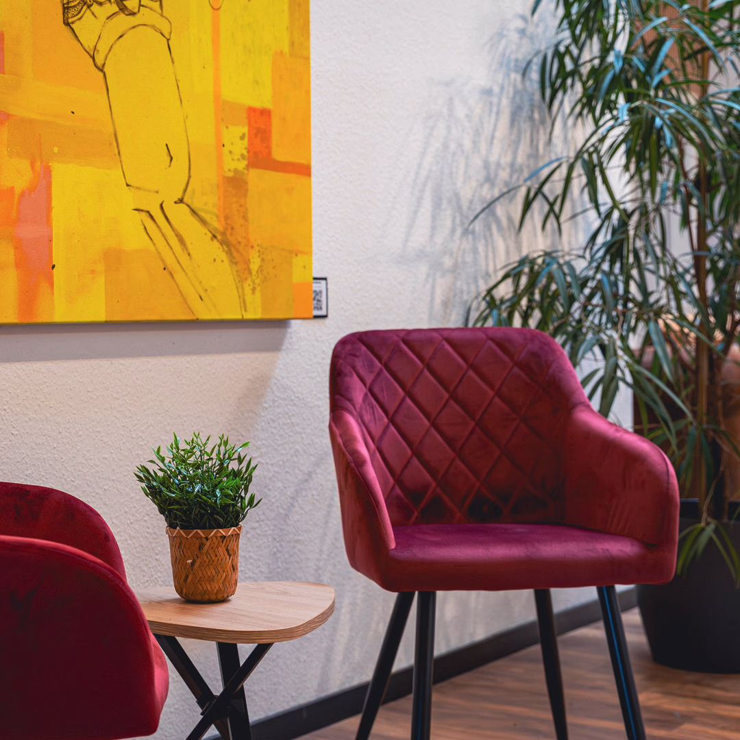 Roter, gepolsterter Stuhl neben einem kleinen Holztisch mit einem Pflanzenkorb, gelbes Kunstwerk und eine große grüne Pflanze im Hintergrund.