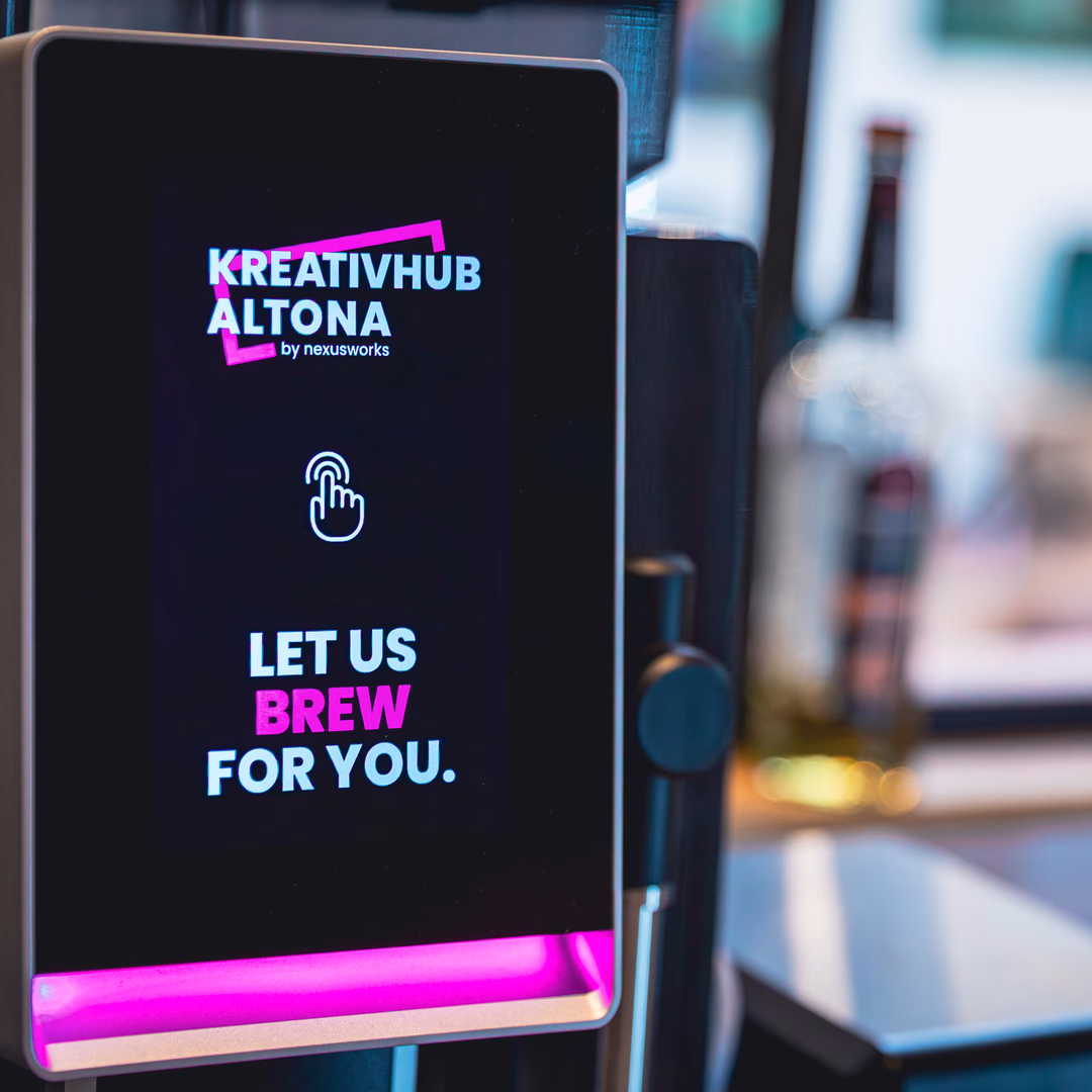 Touchscreen display on a coffee machine with the text ‚Kreativhub Altona by nexusworks‘ and ‚Let us brew for you‘ in a modern font with pink and white colors.