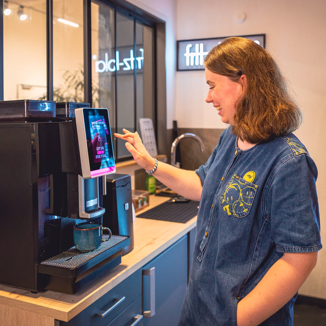 Frau bedient eine moderne Kaffeemaschine mit Touchscreen, während eine Tasse darunter steht.