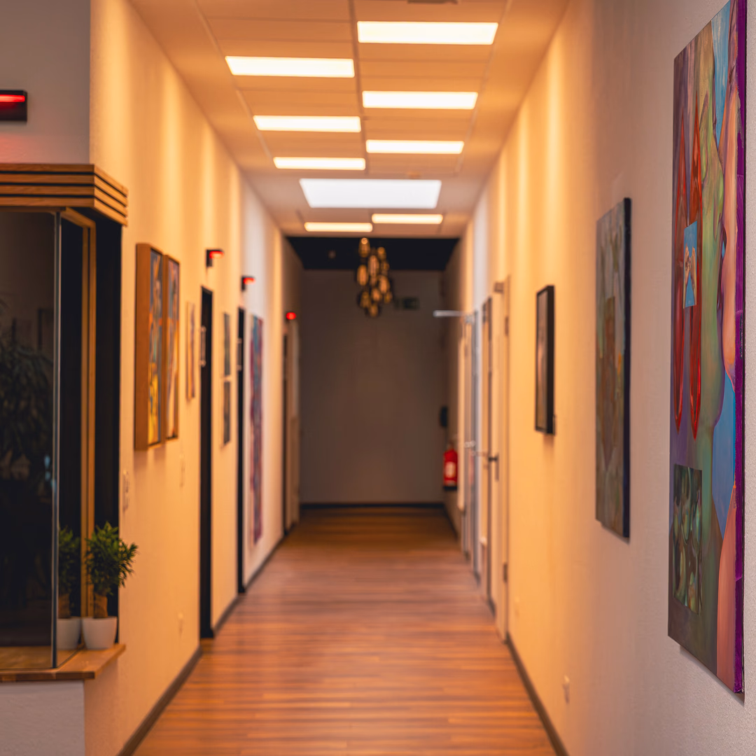 Beleuchteter Flur mit Holzboden, weißen Wänden und bunten Gemälden an den Seitenwänden.