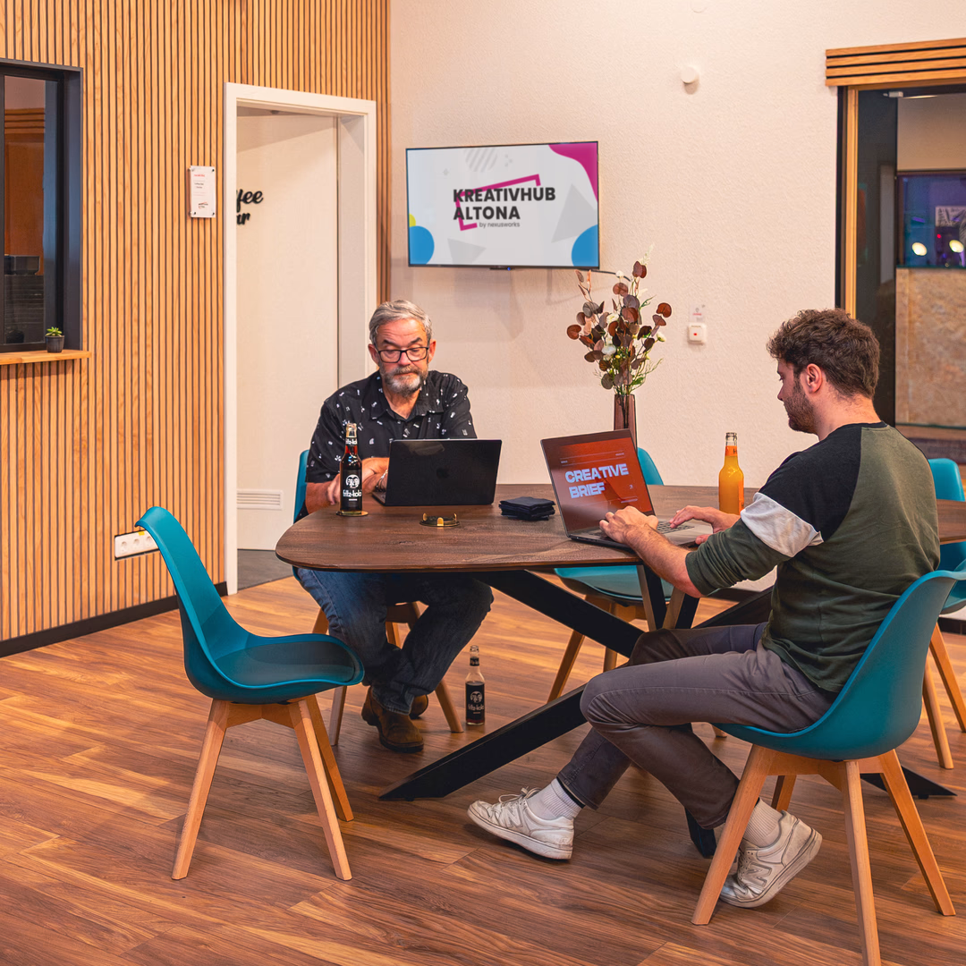 Zwei Männer arbeiten an Laptops an einem Holztisch in einem modernen Büro mit blauen Stühlen und einem Bildschirm mit dem Schriftzug Kreativhub Altona im Hintergrund.