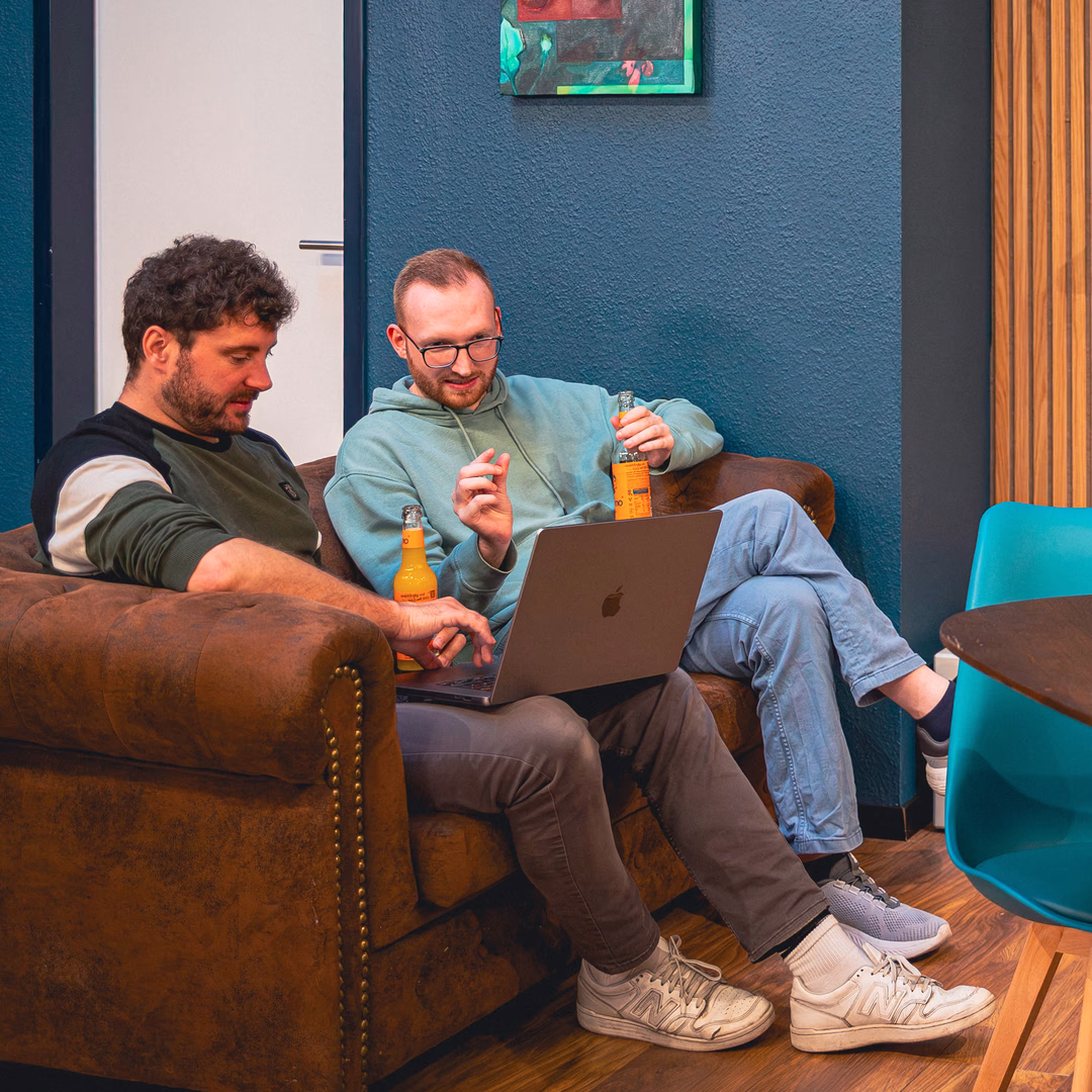 Zwei Männer sitzen auf einem braunen Sofa, arbeiten an einem Laptop und halten jeweils ein Flaschengetränk.