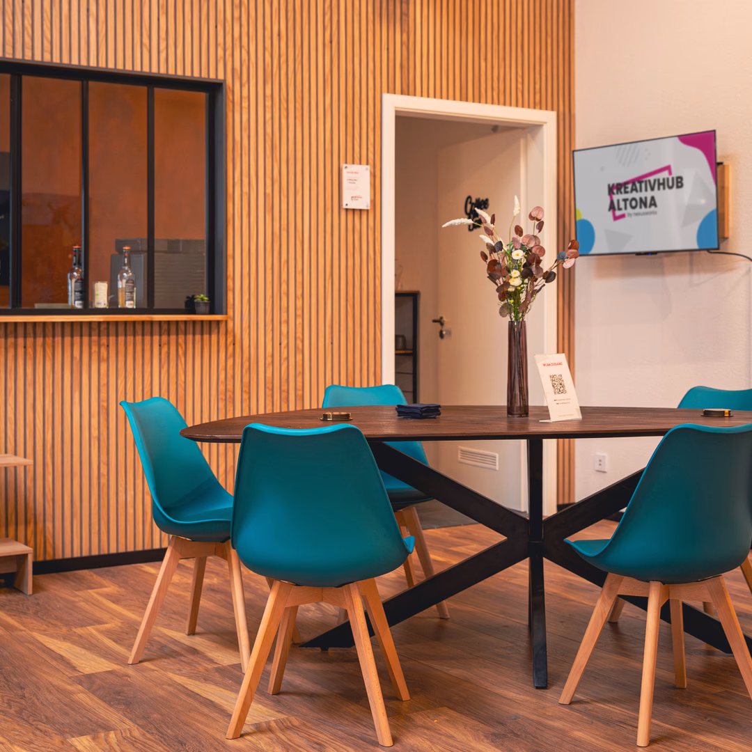 Moderner Besprechungsraum mit einem runden Tisch, blauen Stühlen und einer Vase mit Blumen, im Hintergrund ein Bildschirm mit der Aufschrift Kreativhub Altona.