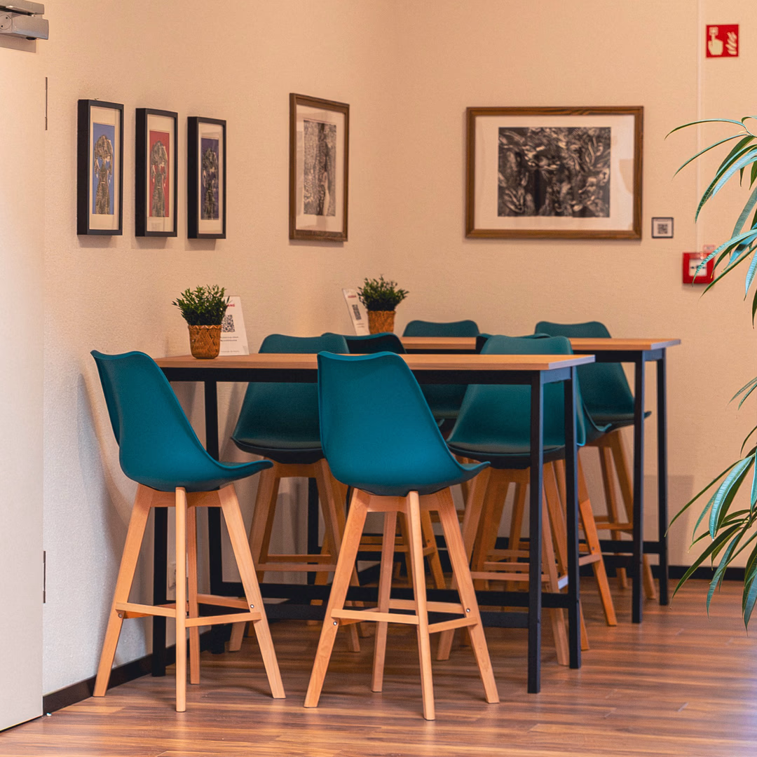 Moderner Sitzbereich mit hohen Tischen, blauen Stühlen, hölzernem Boden, Wandbildern und grünen Pflanzen.