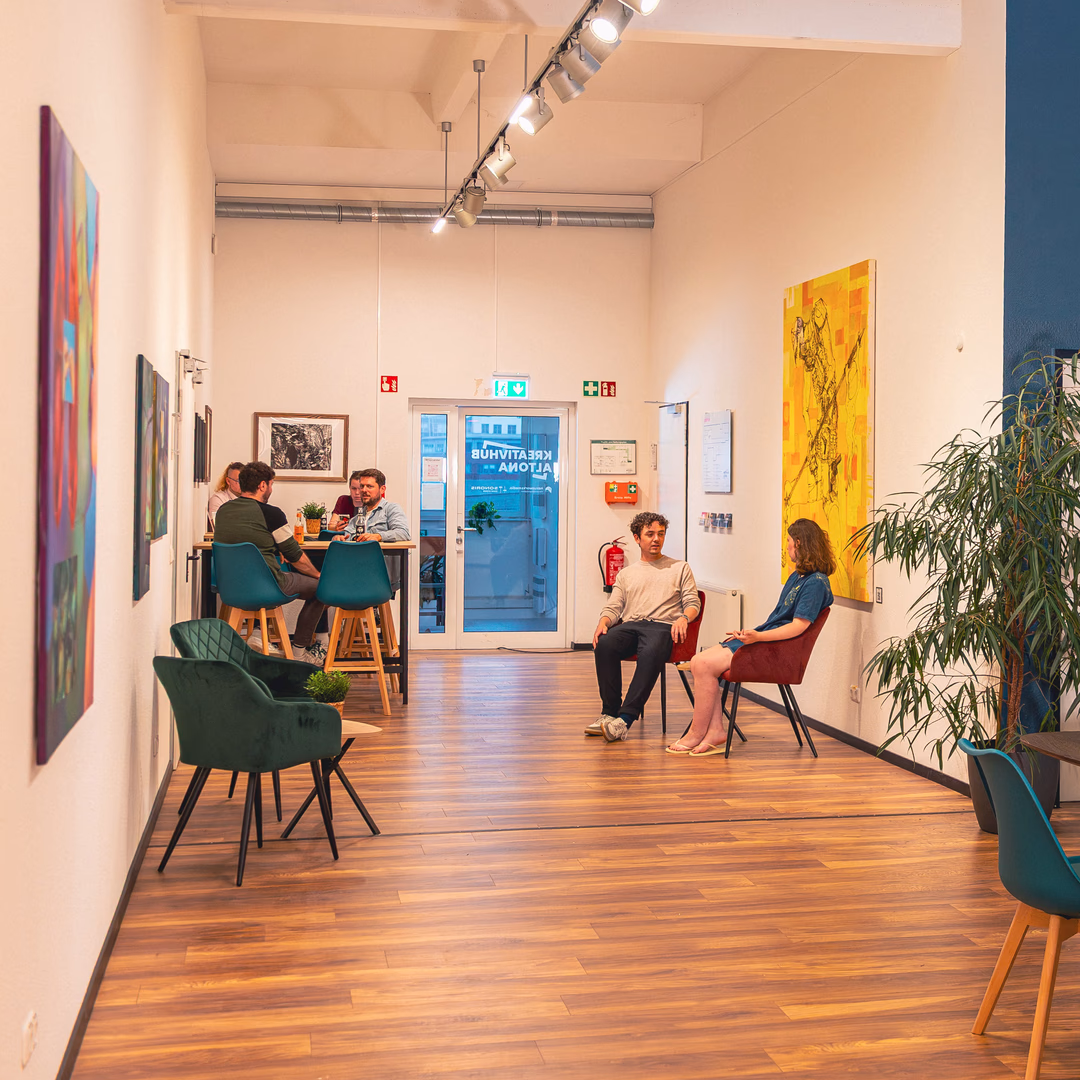 Moderner Gemeinschaftsraum mit Holzboden, mehreren Stühlen, Pflanzen und Menschen, die sich in kleinen Gruppen unterhalten.